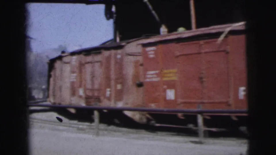 String of rust-red freight boxcars with stencilled markings receding along the tracks under blue sky, distant shed visible, Calcutta goods yard, circa 1962, 8mm colour film.