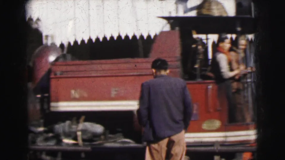 Trackside observer in dark jacket watches the red steam locomotive pass in Calcutta circa 1962; a building with a decorative roofline is visible in the background.