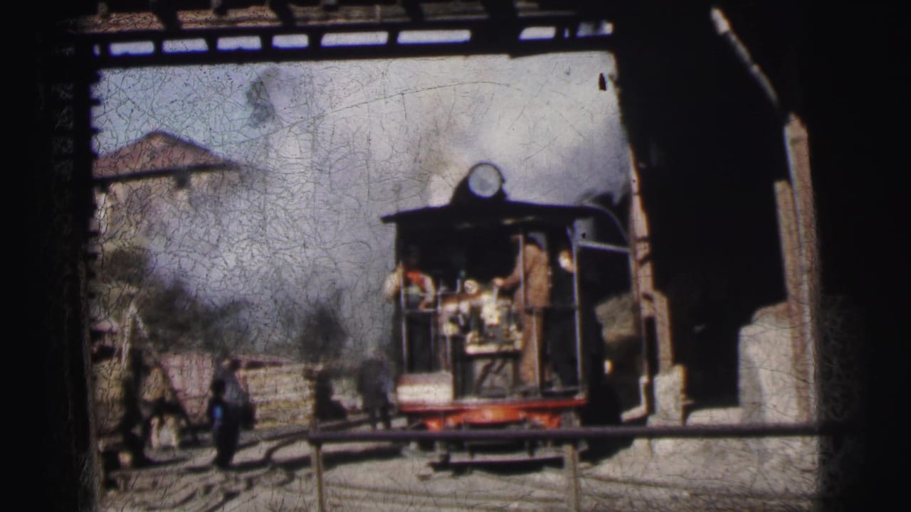 Small narrow-gauge steam locomotive in an industrial yard, workers visible on and around it, building in background