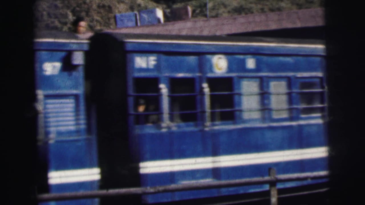 Blue 'NF' railway carriage with white stripe passes through a station in India, circa 1962, passengers visible through barred windows, foliage in background.