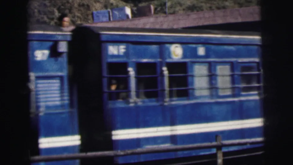 Blue 'NF' railway carriage with white stripe passes through a station in India, circa 1962, passengers visible through barred windows, foliage in background.