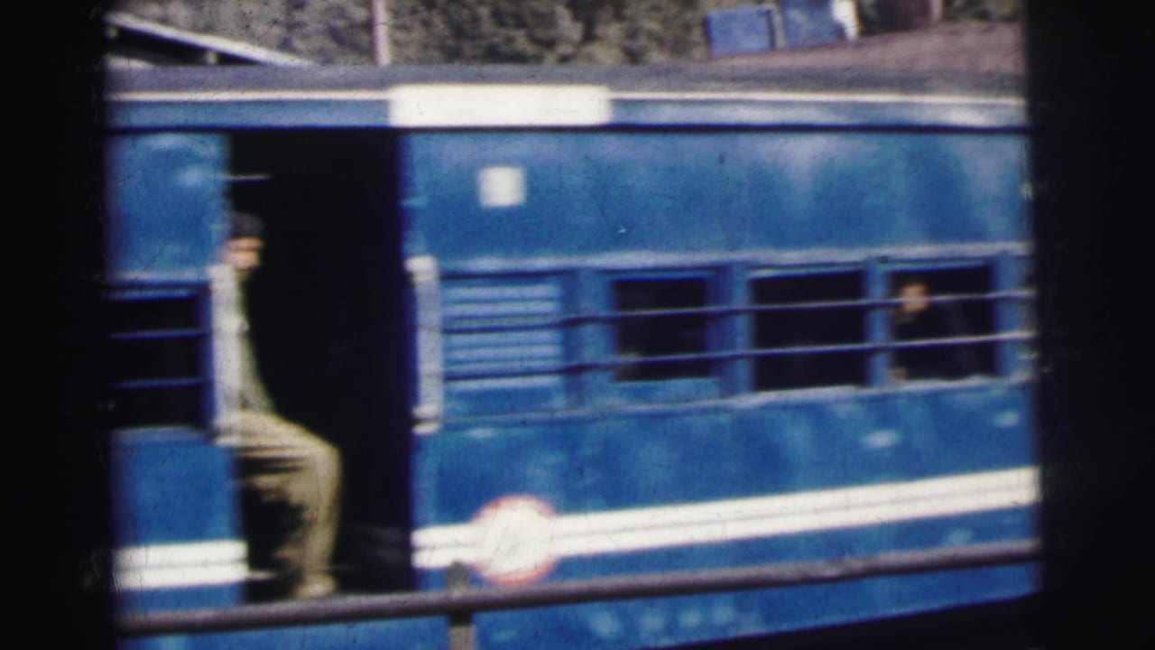 A man in khaki uniform stands in the open doorway of a blue 'NF' carriage with white stripe, passengers visible at barred windows behind him, c. 1962 India.
