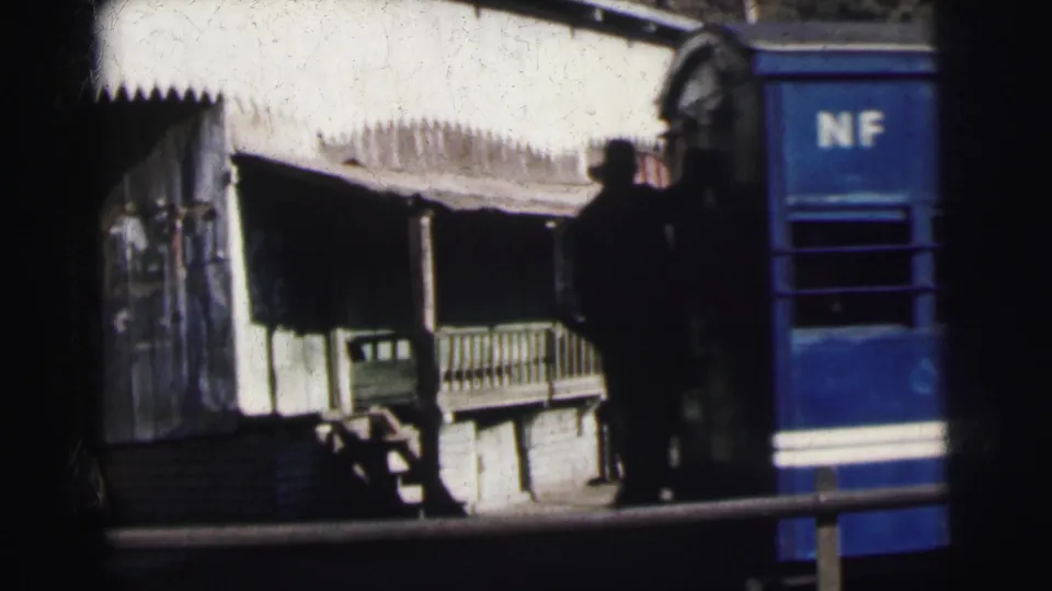 A colonial-era Indian station platform with serrated canopy edge, bench, and standing figures visible, with a blue NF carriage passing, c. 1962.