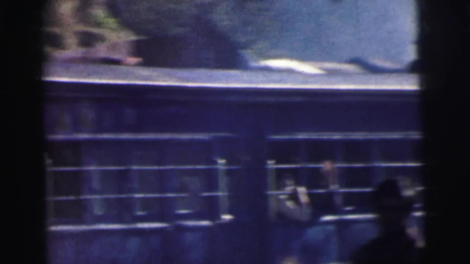 The rear of a blue Indian Railways carriage with white stripe passes the camera, a passenger visible through a barred window, motion blur evident, c. 1962.