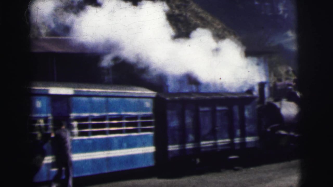 Blue and white passenger train carriages at station platform; steam locomotive billowing white smoke in background; figures on platform.