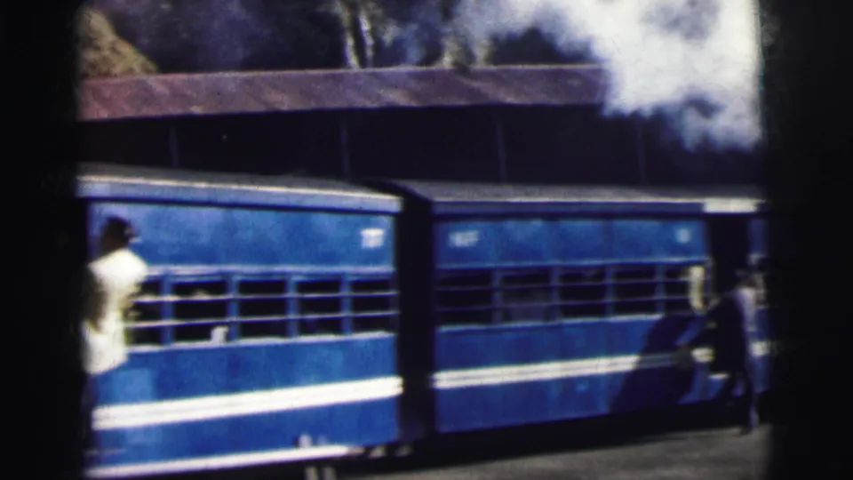 8mm frame, circa 1962 Calcutta — blue and white passenger carriage passing a station platform, with a person standing nearby and a station structure above.