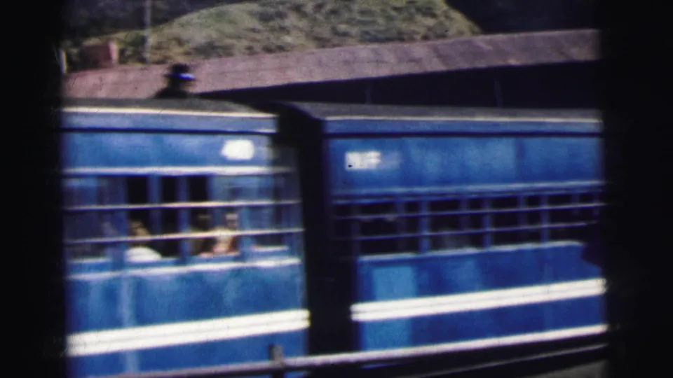8mm film frame, circa 1962 Calcutta, India — passengers lean from open windows of a blue and white painted train carriage passing a station.