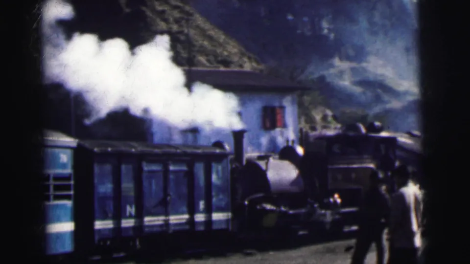 Steam locomotive with heavy steam discharge beside blue carriages at a mountain railway station with hilly landscape, 8mm colour film, India c.1962.