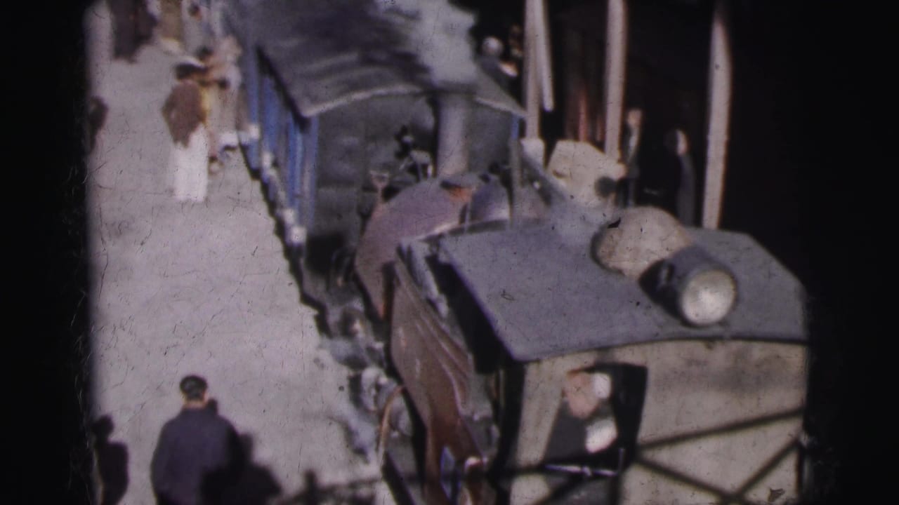 Overhead shot looking along the platform beside blue passenger cars at what appears to be Calcutta railway station, circa 1962, with passengers in period attire visible.