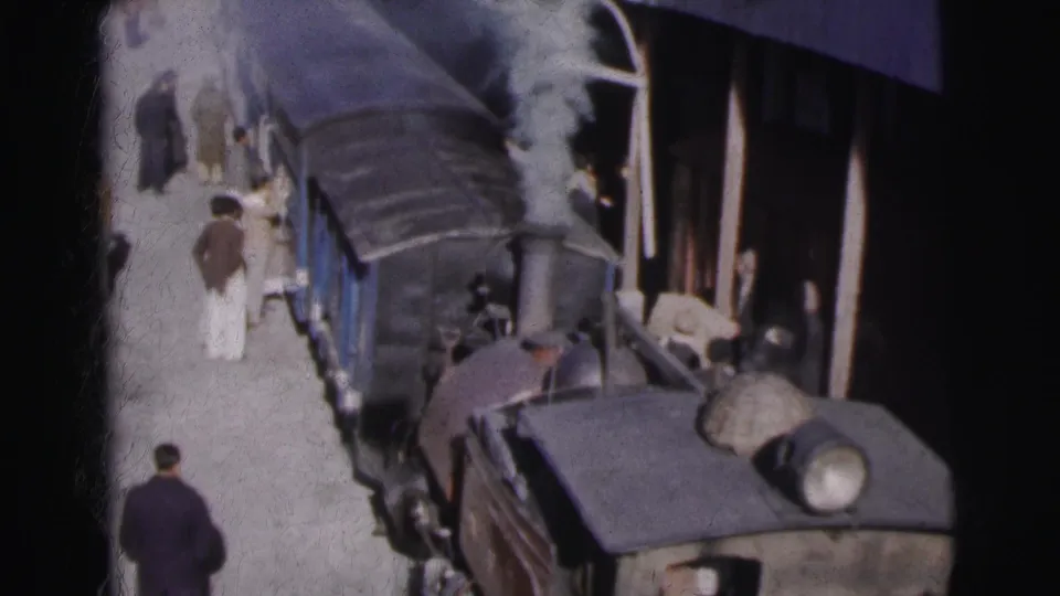 Overhead wide view of a steam locomotive and blue passenger cars stopped at Calcutta station circa 1962, with steam rising and passengers in period dress on the platform.
