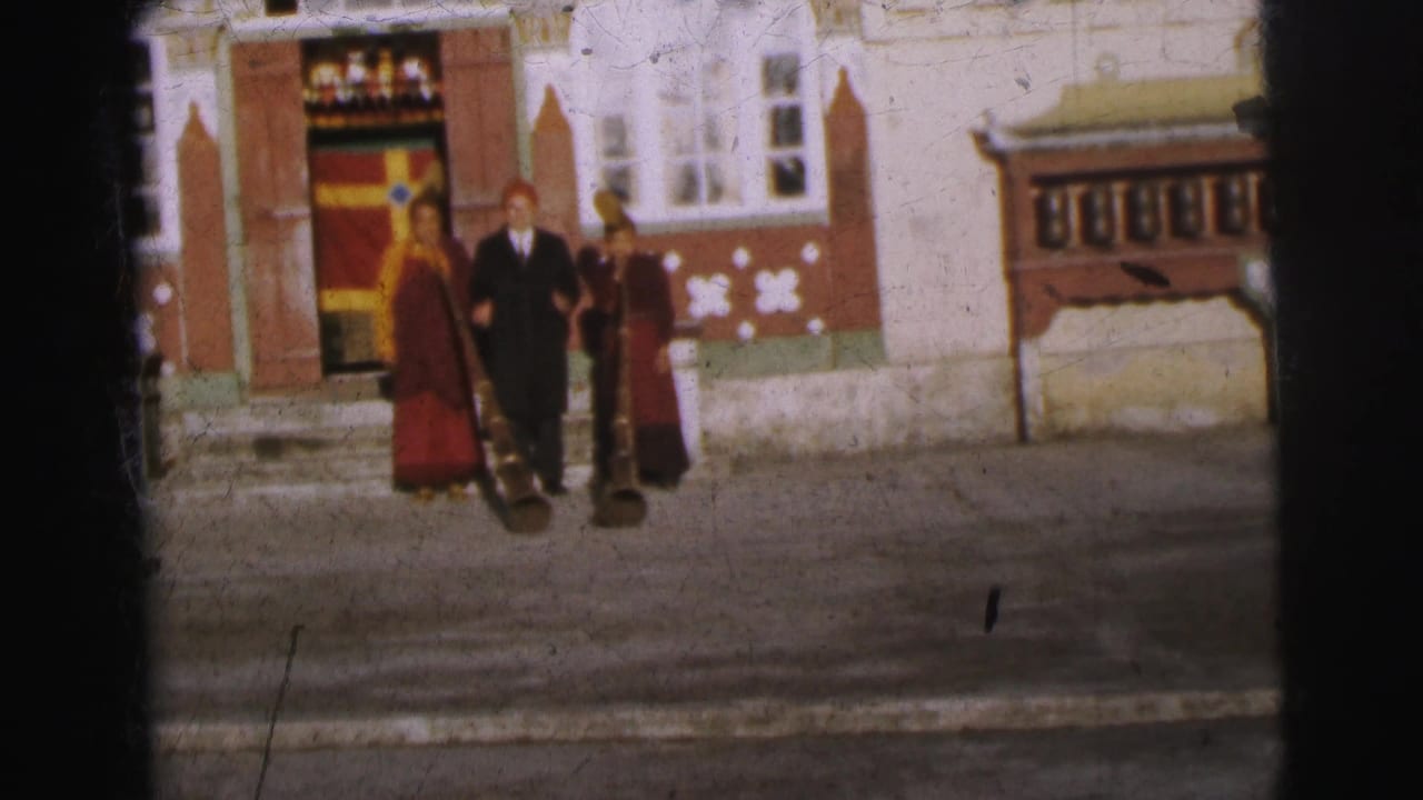 Group of three in red robes and dark suit slightly repositioned before ornate building with red doors and patterned white walls.