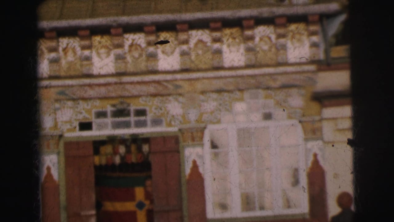 Close-up of decorated building facade showing yellow plasterwork with painted motifs, red banding, white arched stucco elements, wooden shuttered door, and a person partially visible at right edge