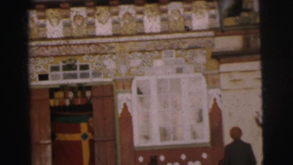 1962 Calcutta, 8mm: facade close-up showing yellow painted ornamental walls, red banding, white arched stucco relief, colorful entrance panels in green, yellow, and red, with a person's silhouette at lower frame edge