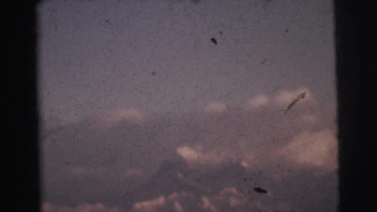 8mm film frame c. 1962 showing a dark mountain outline beneath clouds with multiple film dust particles visible, India.