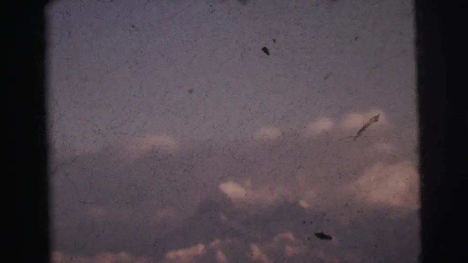 8mm film frame c. 1962 showing a dark mountain outline beneath clouds with multiple film dust particles visible, India.