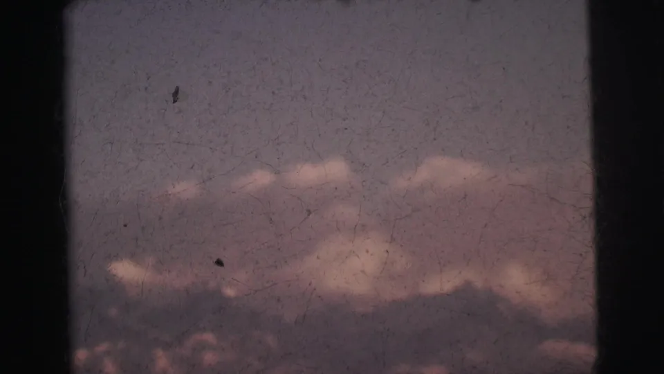 8mm film frame c. 1962 showing a dark mountain ridge below pink and purple twilight clouds, with pronounced grain and emulsion cracking, India.