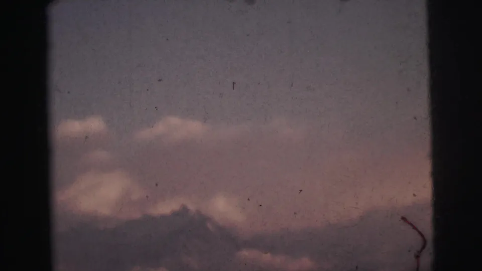 8mm film frame c. 1962 showing a mountain ridge silhouette below pink twilight clouds, with a curved scratch artifact in the lower right corner, India.