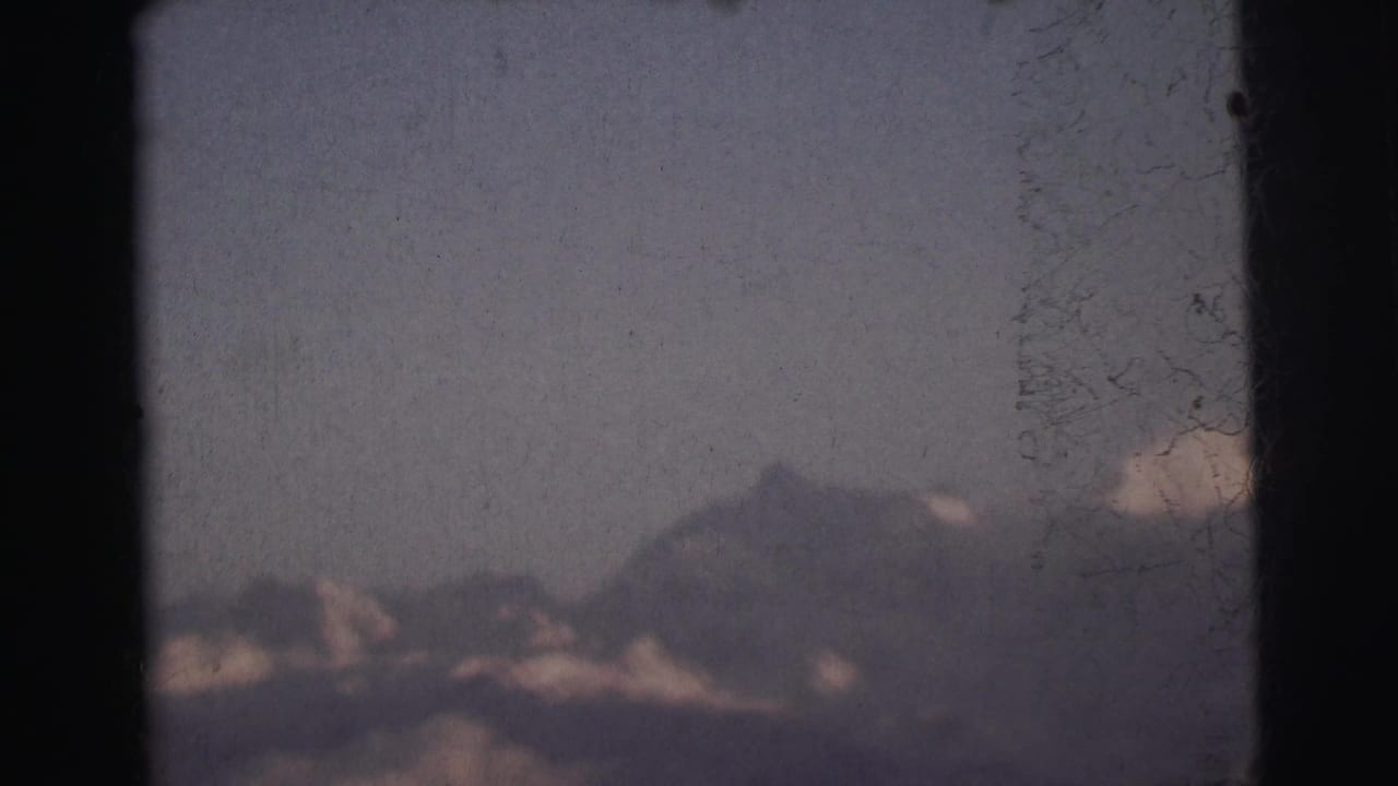 Jagged mountain summit partially veiled in clouds, shot through aged 8mm film with visible scratches and grain.