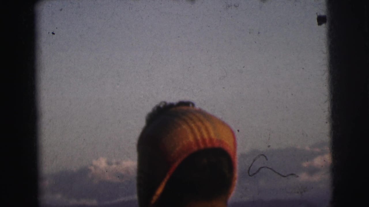 Rear view of passenger in striped cap at train window, distant mountains and overcast sky behind, 8mm film.