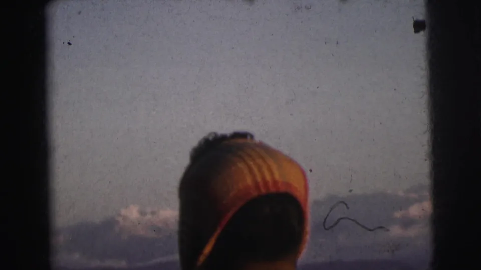 Same traveler in striped knitted cap seen from behind at a train window, hazy mountain range in background, 8mm home movie, India, 1962.