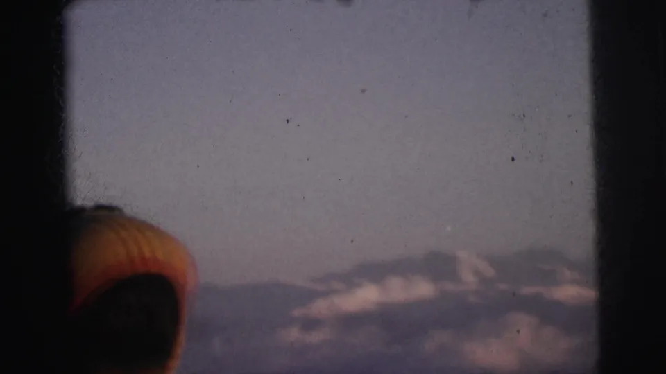Camera reveals snow-capped mountain peaks and wide pale sky through a train window; passenger's cap remains visible at frame edge, 8mm film, India, 1962.