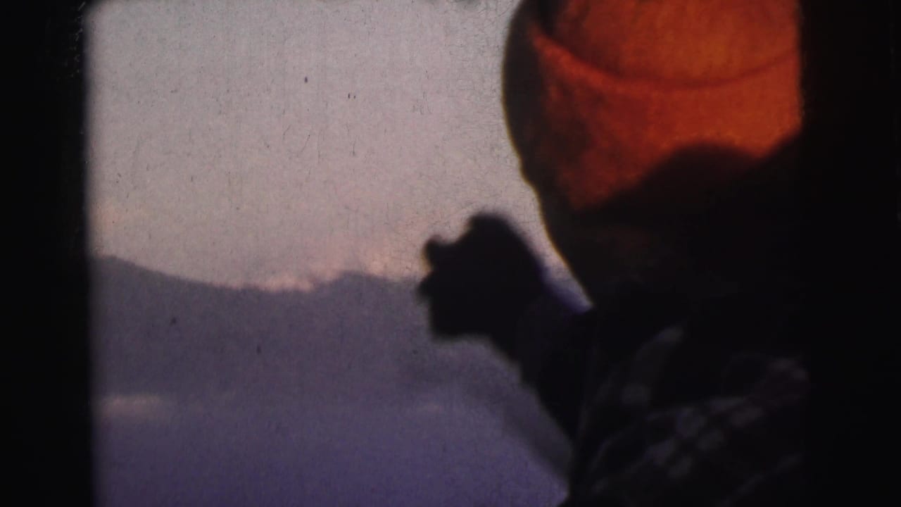 Person in orange-red knit hat raises hand toward a misty mountain horizon, 8mm home movie footage, India, circa 1962.