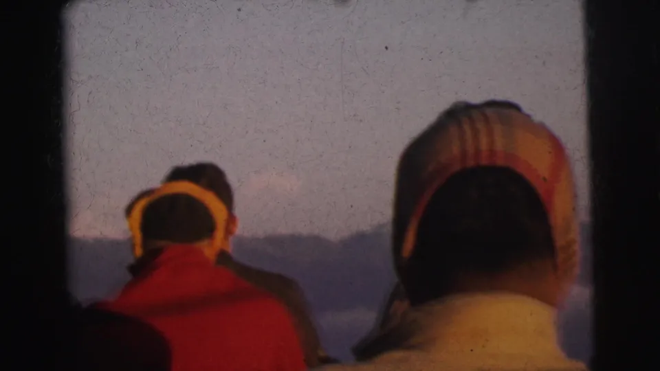 Close group of figures in winter hats face a distant mountain ridge in 8mm home movie footage, India, circa 1962.