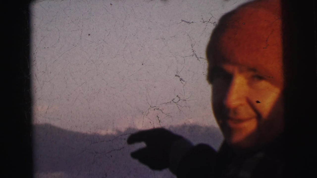 Man in red knit beanie gestures toward a pale mountain landscape in close-up, 8mm film.