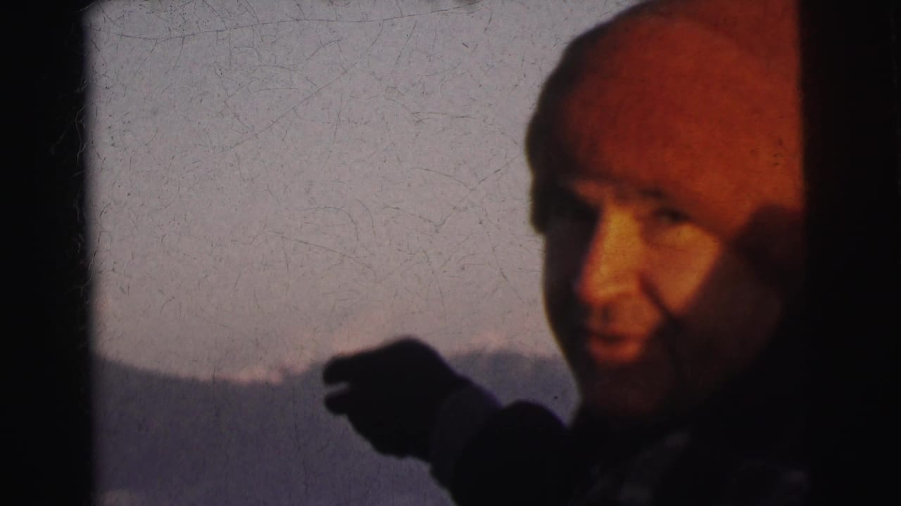 Close-up of a man in a red knit beanie with a gloved hand gesturing toward a hazy mountain landscape, 1962 8mm home movie footage from India.