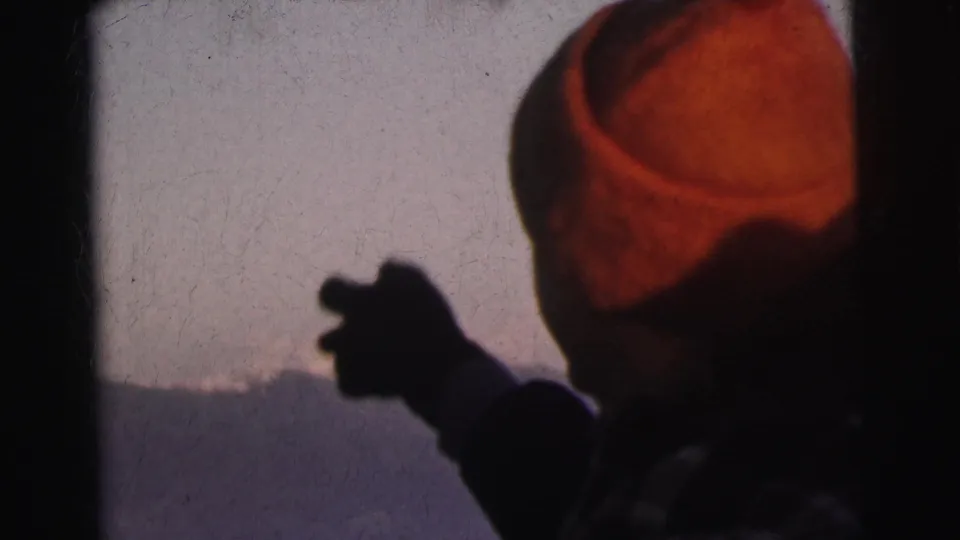 Rear three-quarter view of a man in a red knit beanie gesturing toward a snowy mountain landscape, 1962 8mm home movie footage from India.