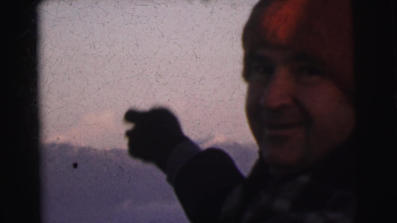 Man in red wool beanie and dark coat gesturing toward camera, distant mountain range behind him, 8mm footage from India circa 1962