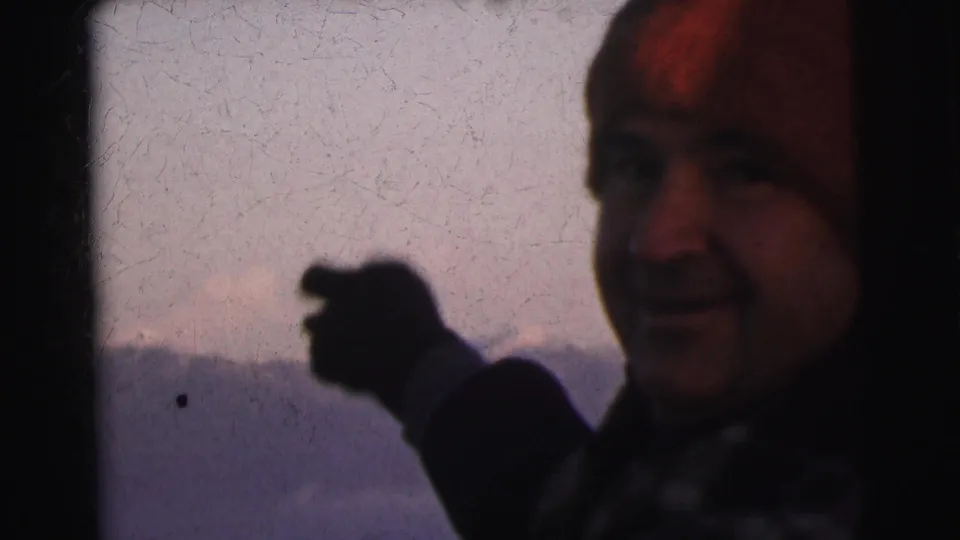 Man in red wool beanie and dark coat gesturing toward camera, distant mountain range behind him, 8mm footage from India circa 1962