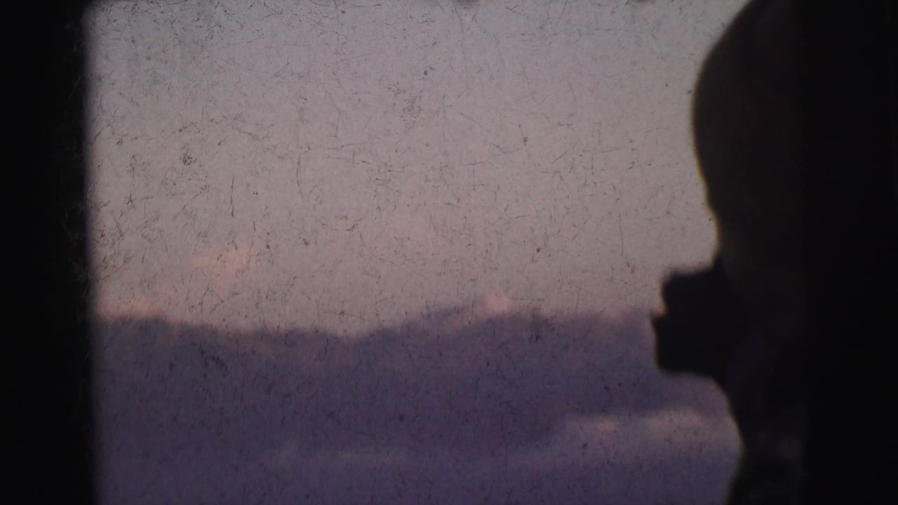 Figure in near-silhouette with hand raised against pale lavender-blue sky and mountain silhouette, 8mm footage circa 1962