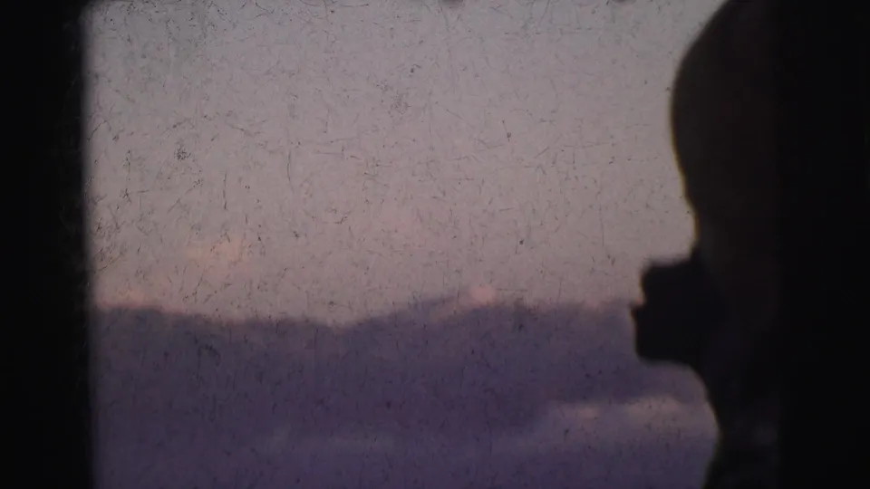 Figure in near-silhouette with hand raised against pale lavender-blue sky and mountain silhouette, 8mm footage circa 1962
