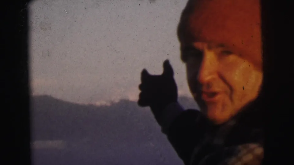 Man in red beanie and dark coat speaking to camera while gesturing, mountain range in background, 8mm home movie circa 1962 India