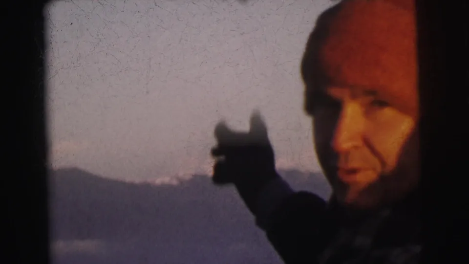 Close framing of man in red beanie gesturing toward landscape, pale sky and mountain range behind, 8mm footage from India circa 1962
