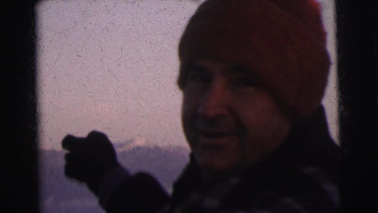 Close-up of a man in a red beanie and dark coat gesturing with dark-gloved hands; a mountain ridgeline is visible in a soft purple background.