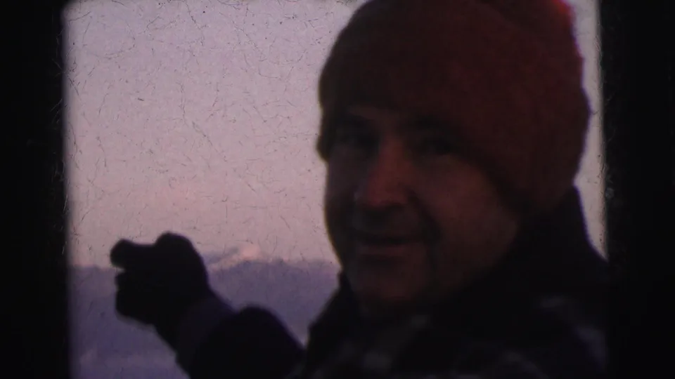 Man in red knit beanie and dark winter coat gesturing with dark gloves toward unseen peaks; mountain ridgeline in soft dusk light, 8mm film circa 1962.