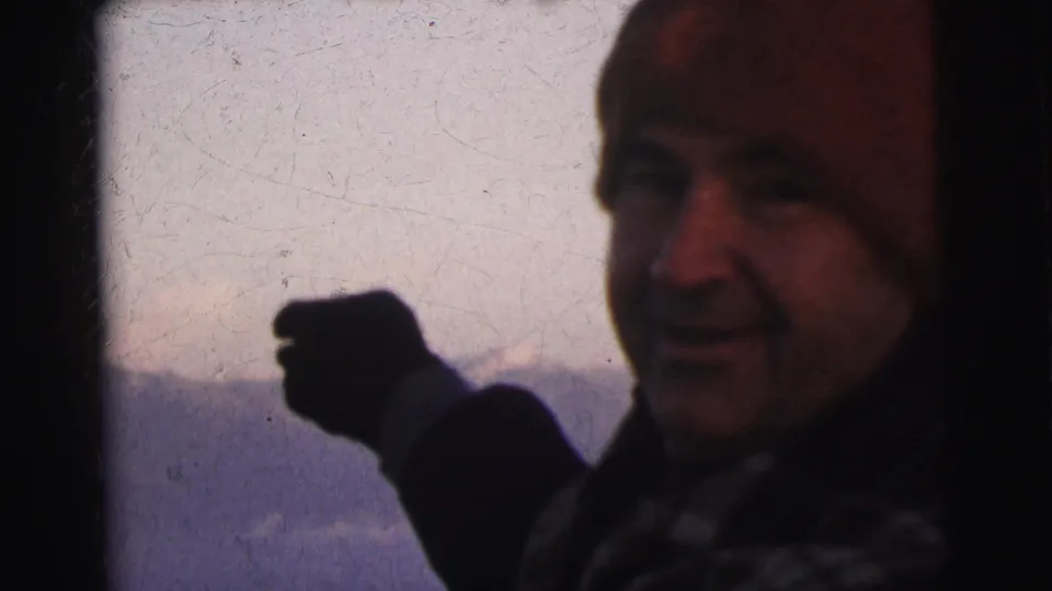 Man raising a dark-gloved hand with snow-capped mountains faintly visible behind him; 8mm footage with notable grain and scratch texture, circa 1962.