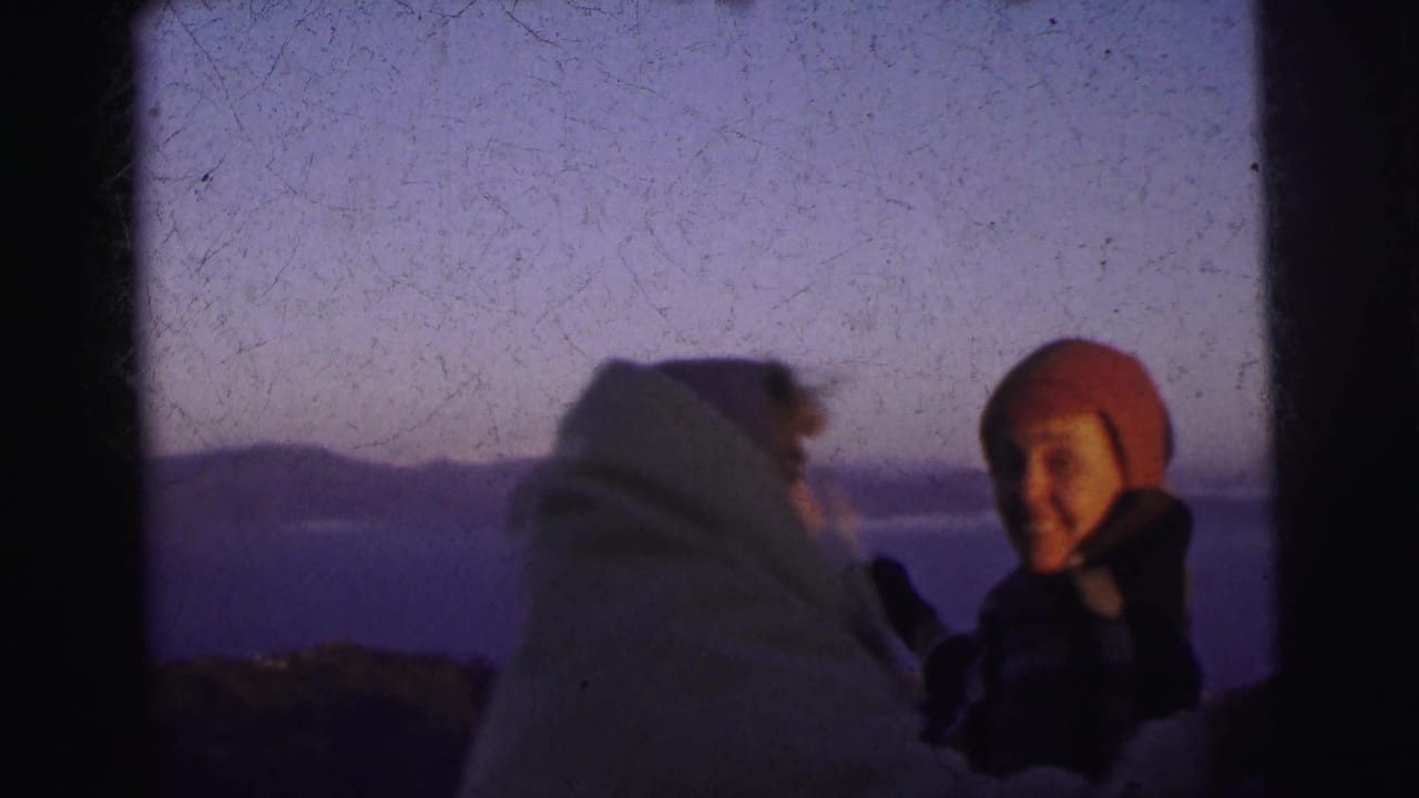 Two adults at dusk in footage described as Calcutta, India, 1962; one wears a red wool beanie and smiles at the camera, the other faces away wrapped in a pale blanket.