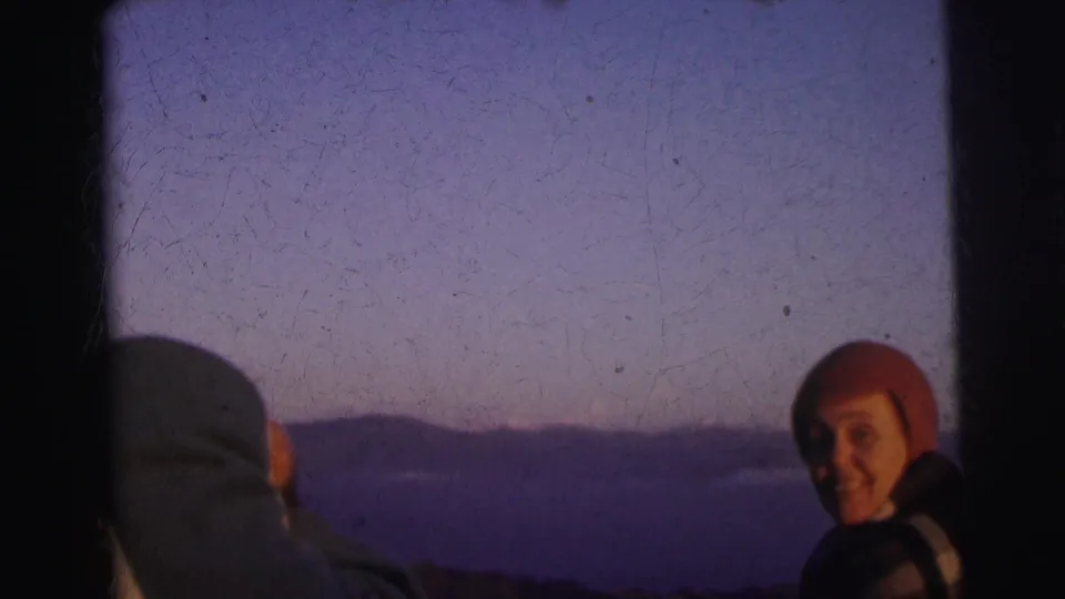 Smiling adult in red wool beanie at twilight, second adult seen from behind at left; 8mm footage described as Calcutta, India, 1962.