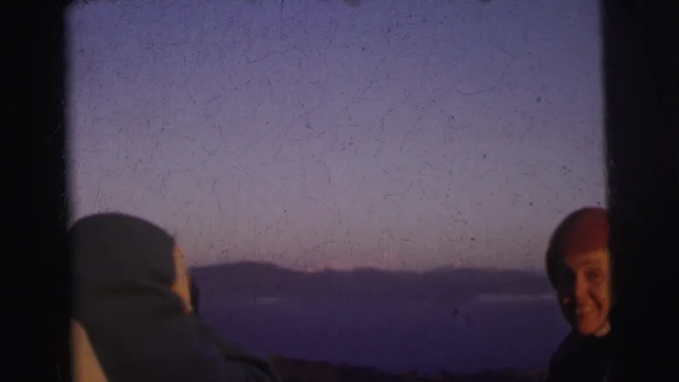 Close-medium view of smiling adult in red beanie outdoors at dusk, second figure in pale wrap at lower left; 8mm footage described as Calcutta, India, 1962.