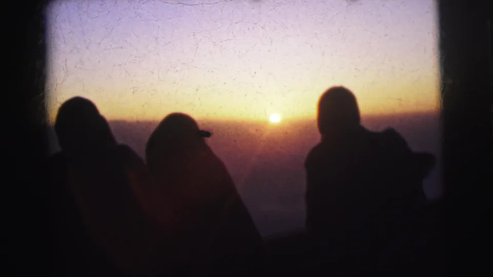 Silhouetted figures, one in a cap, watch an orange sunset from a high vantage point; emulsion crazing spreads across the upper sky, consistent with aged 8mm stock; metadata places this in Calcutta, 1962.