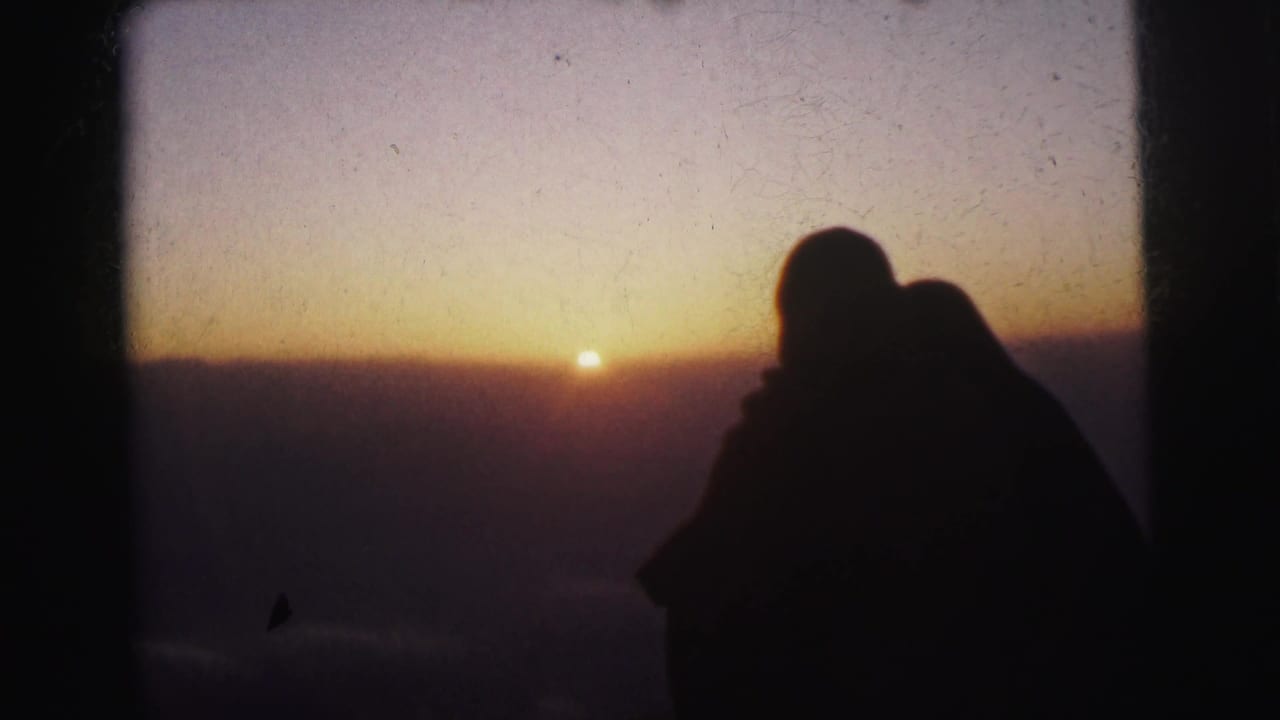 In supported 1962 Calcutta home-movie footage, two adults appear in silhouette near a window or frame edge as the sun sits low on an orange-purple horizon.