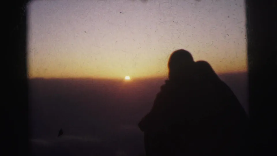 In supported 1962 Calcutta home-movie footage, two adults appear in silhouette near a window or frame edge as the sun sits low on an orange-purple horizon.