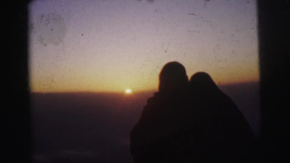 The couple's dark outline is held against a golden sunset in the supported Calcutta 1962 home-movie scene, with soft color and visible 8mm wear across the frame.