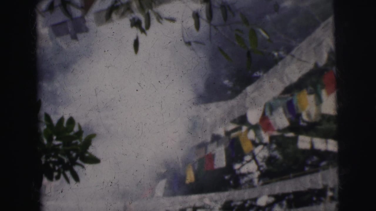Colorful cloth prayer flags strung on a line between white railings, tree foliage at upper left, hazy sky, 8mm film grain and surface scratches visible.