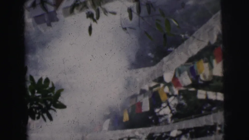 Colorful prayer flags on a string near white railing structure, tree foliage overhead, 8mm footage from Calcutta, India, circa 1962.