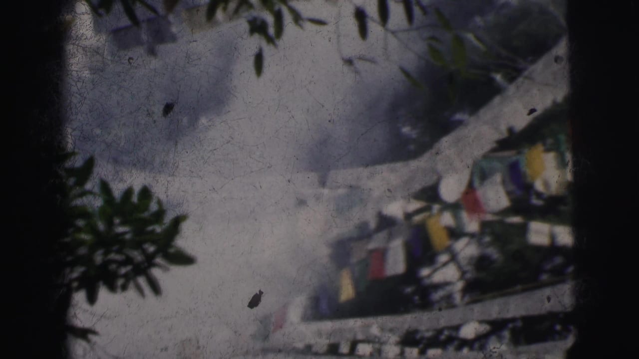 Multicolored prayer flags strung between white structures, tree branches at upper right, hazy sky, 8mm film grain and scratches visible.