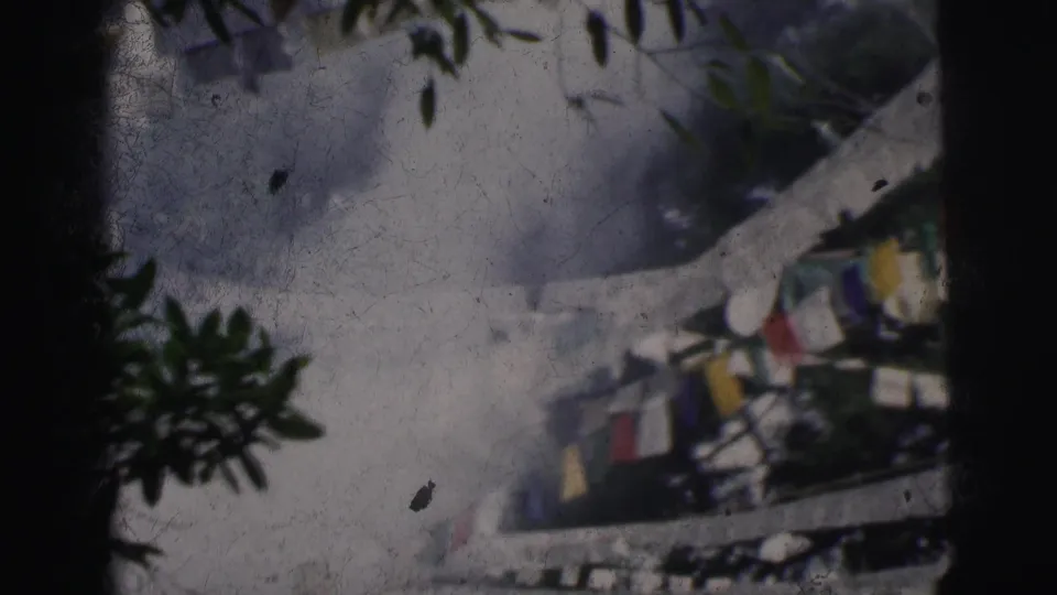 Multicolored cloth prayer flags near a white railing structure, tree branches visible at top, 8mm footage from Calcutta, India, circa 1962.